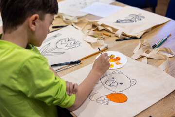 Child painting on a canvas tote bag during a creative art therapy workshop. Eco-friendly kids'...