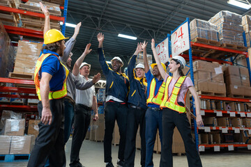 Diverse group of warehouse workers wearing safety gear celebrates with raised hands for teamwork,...