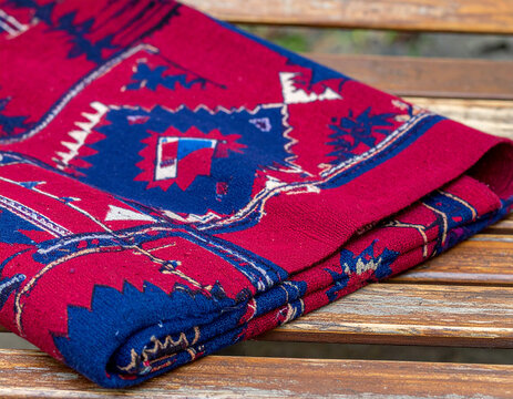 A close-up of a handmade Pakistani ajrak shawl with deep red and indigo geometric motifs spread across a wooden bench.