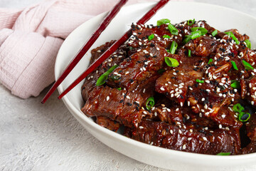 Crispy beef, with green onions and sesame seeds, in a bowl with chopsticks, on a textured surface, on a light background, no people