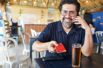 Smiling man paying by credit card while talking on phone in a Caribbean bar