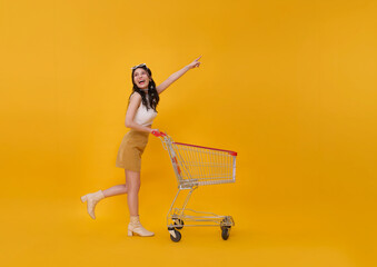 Full length happy excited Asian teen woman pushing empty shopping cart or shopping trolley pointing finger promotion supermarket isolated on yellow background. © NaMong Productions