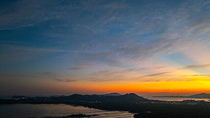 A stunning panoramic view of a coastal city at sunset at Phuket city, showcasing vibrant hues of orange and blue in the sky, with mountains, and urban lights creating a harmonious blend of natural.