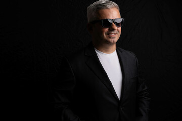 Obraz premium Happy confident man wearing sunglasses posing against black background. Low key lighting portrait, medium shot, dark and moody atmosphere