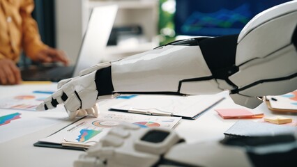 Robotic hand engaging with data charts in an office showcases the integration of technology into business analysis and the advancement of artificial intelligence.Noogenesis