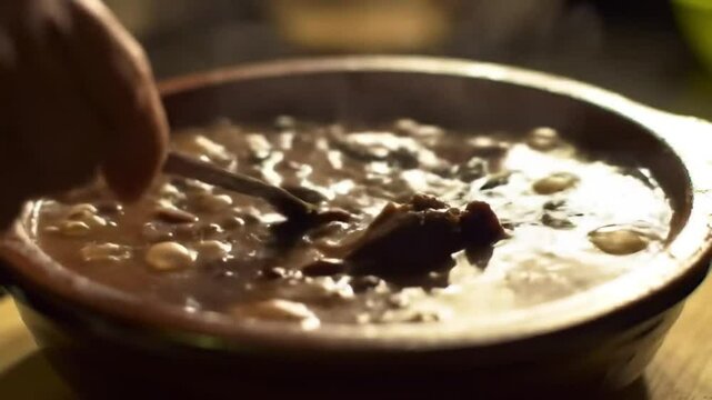 Close-up video of traditional feijoada in a pot, steam rising, a spoon slowly stirring the rich, thick stew, showcasing hearty ingredients, rustic kitchen atmosphere.

