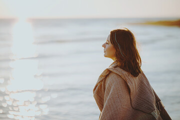 A woman in a blanket on the seashore breathes freely and looks thoughtfully into the distance