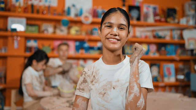 Happy caucasian girl pose at camera while diverse children modeling clay behind. Cute student wearing dirty shirt while looking at camera at workshop in art lesson. Blurring background. Edification.
