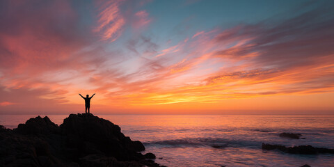 Silhouette of person with arms raised, standing on coastal rock against vibrant sunset sky and ocean, conveying freedom, peace, and achievement