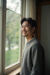 Smiling Asian Man in Casual Sweater Looks Out Large Window Towards Blurry Park View, Relaxed Home Lifestyle