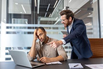 A frustrated manager confronts a stressed employee in an office setting, pointing at a laptop. The...