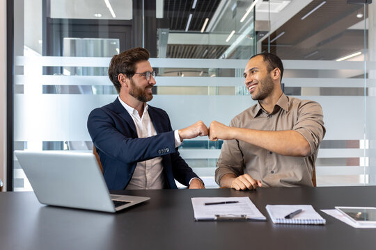 Two men, likely colleagues, bump fists in a modern office environment. A laptop and documents are on the table, indicating a business meeting or discussion.