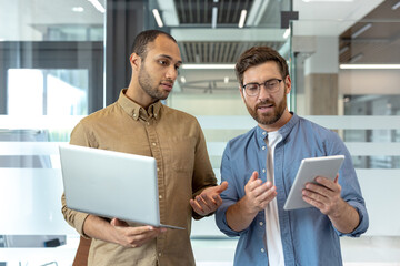 Two professional men collaborate in a modern office, using a laptop and tablet for discussion. They...
