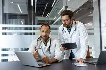Two doctors in white coats are discussing something on a laptop, a clipboard, and a tablet in a...