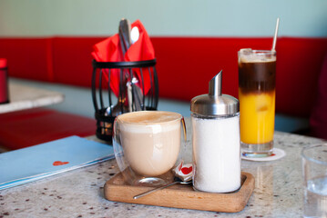 a clear glass glass with coffee, a jar of sugar, red napkins, and a coffee tonic are on the table in the cafe
