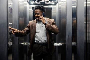 Smiling interracial elegant man typing number in elevator while having phone call.