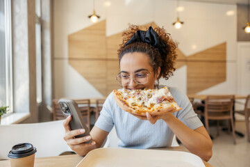 Young multiracial woman sitting in fast food restaurant with cellphone and eating pizza.