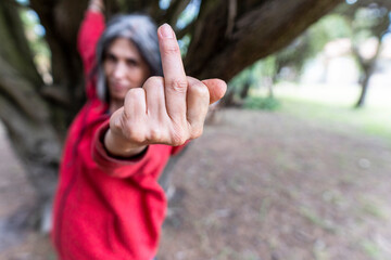 Grey hair woman expresses defiance at camera while standing near a tree in a natural setting.