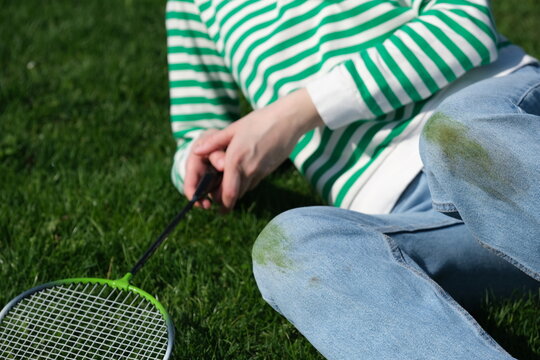A person sitting on the grass with kneeled jeans, holding a badminton racket. The sunny weather playful afternoon activity. Concept of daily stains on clothes.
