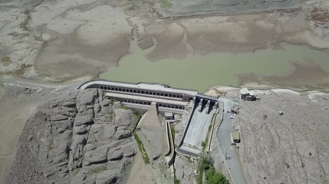 Drone shot directly above the dam capturing the greenish water in the upper reservoir and the cascading slope of the dam wall with multiple arched levels, offering contrast between nature and engineer