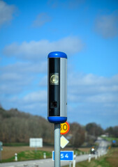 A speed camera stands tall beside a road, capturing the speed of passing vehicles. The device contrasts with the blue sky and hints at a peaceful rural setting