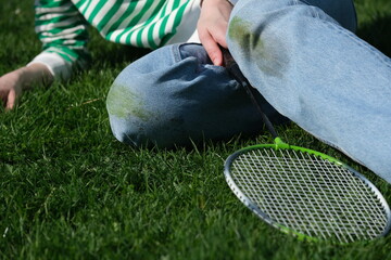 A person sitting on the grass with kneeled jeans, holding a badminton racket. The sunny weather...