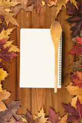 Top view of a blank spiral notebook with a wooden spoon on a rustic wood table, surrounded by colorful autumn leaves. Perfect for seasonal recipes, blogs, or fall-themed culinary designs.