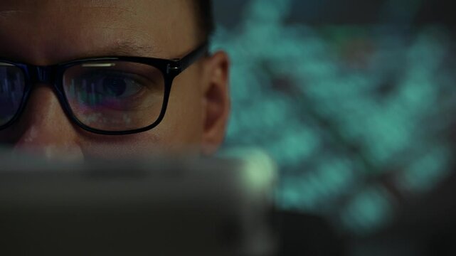 An individual with glasses is intently looking at a laptop screen in a low-light setting. The soft glow of the screen reflects their concentration during late-night work