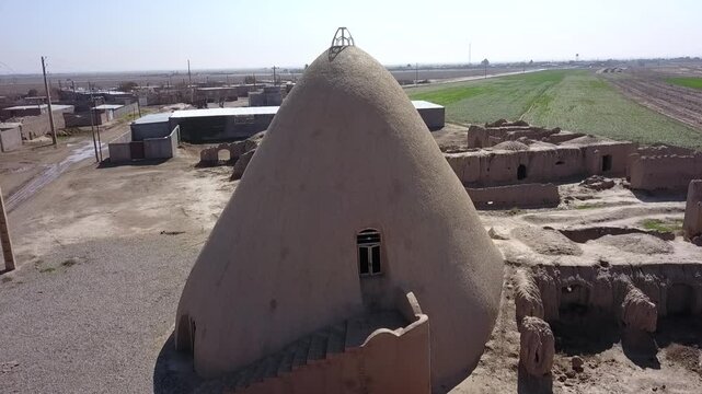 Drone flight above a traditional Persian yakhchal with a large conical dome and thick adobe walls in khorasan iran, surrounded by ruins of a desert village and green farmlands in the distance