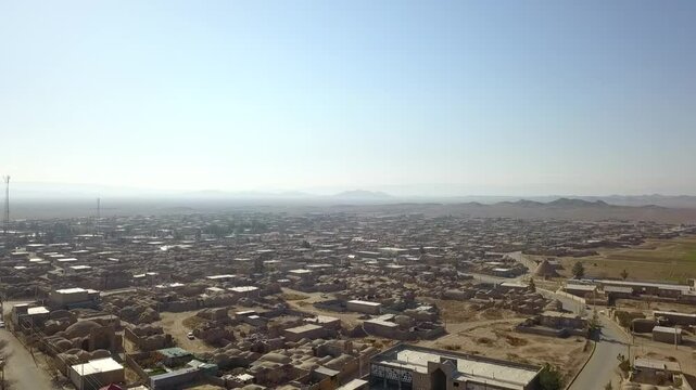 Tilted aerial view of a large Iranian town built on flat terrain with closely arranged buildings and sparse vegetation, surrounded by distant desert fields. The frame shows adaptation to land