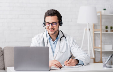 Smiling doctor in white coat with stethoscope and headphones, takes notes in notebook looks in...
