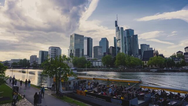 Frankfurt's skyline with Commerzbank Tower and Main Tower, Main River promenade, pedestrians, cyclists, and a lively docked boat gathering.