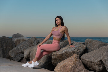 woman is confidently seated on large rocks beach during sunset wearing pink workout attire. ocean waves gently lap against shore as sun sets in background.