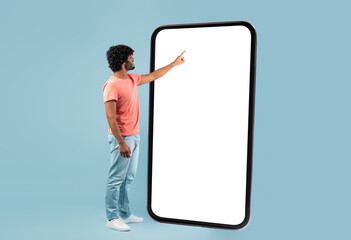 Stylish curly young hindu guy in casual outift touching huge smartphone white empty screen over...