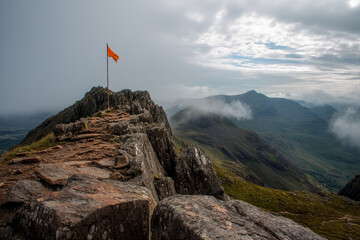 Naklejka premium Orange flag atop rocky mountain peak, overlooking misty valley. Symbol of achievement, exploration, and reaching the summit