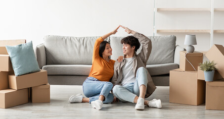 Happy moving day concept. Positive young Asian couple relocating to their own home, making house roof gesture together, sitting on floor among cardboard boxes, copy space. Panorama