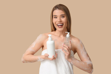 Happy young woman in towel with bottles of shower gel and foam on beige background