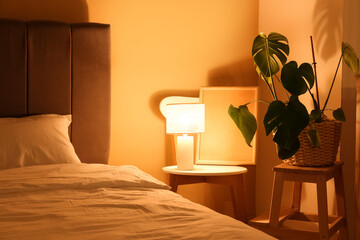 Interior of night bedroom with big bed, houseplant and glowing lamp on chair near yellow wall
