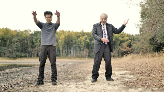 In a spring park with a pond and bamboo grove, a man in his 70s in a suit and down jacket and a man in his 30s practice tai chi. Surrounded by fresh greenery, they move slowly in the calm spring air.