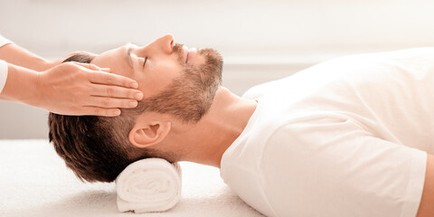 Bearded man enjoying relaxing head massage at modern male spa, side view. Middle aged man laying on massage table with closed eyes, unrecognizable masseuse rubbing his temples, stress and pain relief