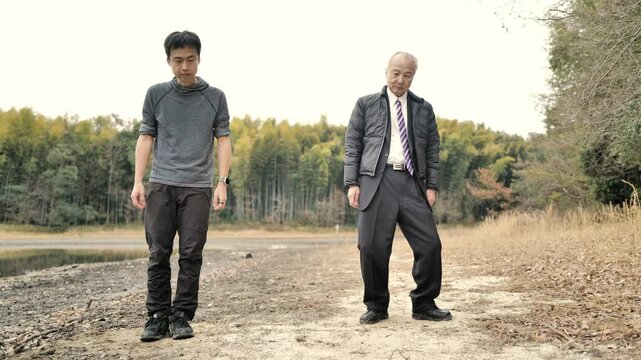 In a spring park with a pond and bamboo grove, a man in his 70s in a suit and down jacket and a man in his 30s practice tai chi. Surrounded by fresh greenery, they move slowly in the calm spring air.
