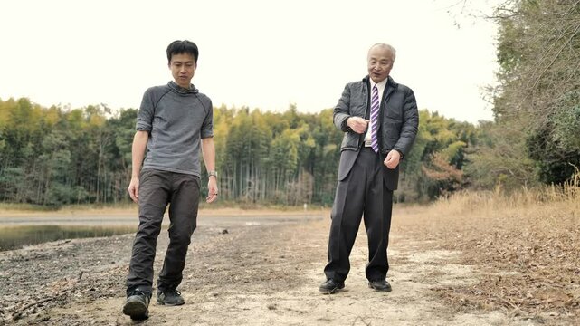 In a spring park with a pond and bamboo grove, a man in his 70s in a suit and down jacket and a man in his 30s practice tai chi. Surrounded by fresh greenery, they move slowly in the calm spring air.