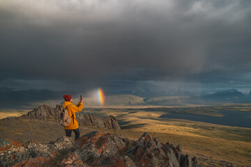 A traveler in bright clothes stands on the edge of a mountain as a rainbow appears in the sky. Golden hour in the mountains with a view of the rainbow and the valley illuminated by sunset.