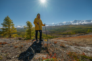 A man in bright clothes stands on the hill of a mountain, the sun is shining above his head, and behind him there are snowy mountains and a green forest. The action takes place in autumn in the mounta
