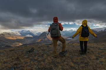 Urist travelers in bright clothes stand on a hill during the golden hour, in front of them there is a view of snowy mountains and valleys with lakes.