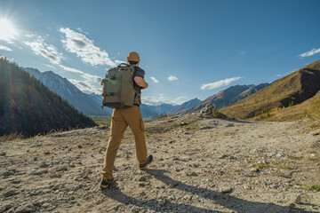 A human traveler with a backpack on his back walks along a mountain path, holding his hands by the straps of the backpack. In front of it there is a view of the mountains and the blue sky.
