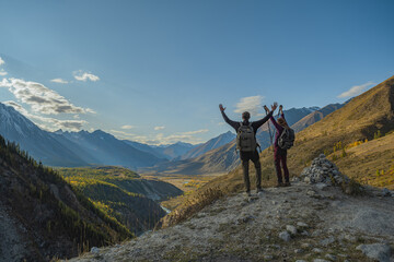 Two travelers with backpacks are standing next to an archaeological site in the mountains, overlooking a large valley and raising their hands to the top.