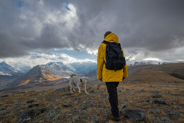 A traveler in bright clothes with a backpack on his back, walks against the background of snowy mountains during the golden hour with his pet. A man and a dog in the mountains.