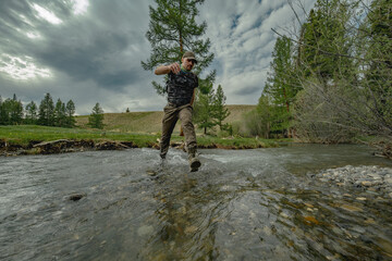 A traveler runs across a wide river against the background of a green forest. A man in boots runs through the water. Trekking clothes for travel.