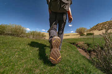 A traveler with a backpack on his back, walking through the green grass, defiantly showing the sole of his shoe. But the background is a sandy dune, bushes and a blue sky.